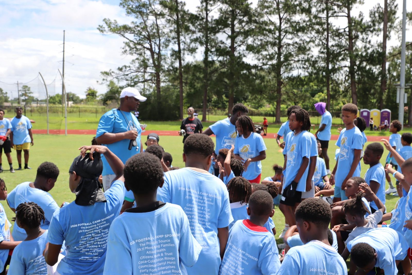 Campers in light blue 2026 Football Camp T-shirts gathered on a grass field around an adult in a matching shirt and white cap holding a megaphone.